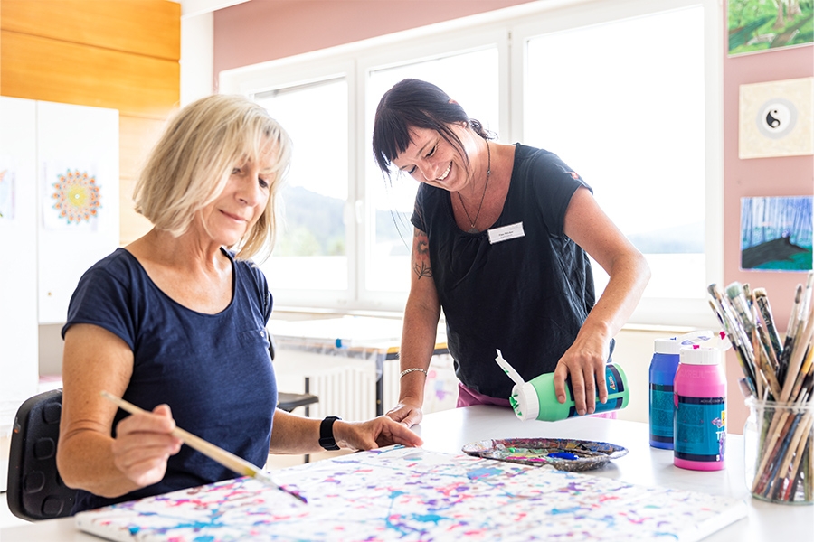 Eine Therapeutin füllt die Farbe einer Patientin nach, die gerade ein Bild mit einem Pinsel malt.