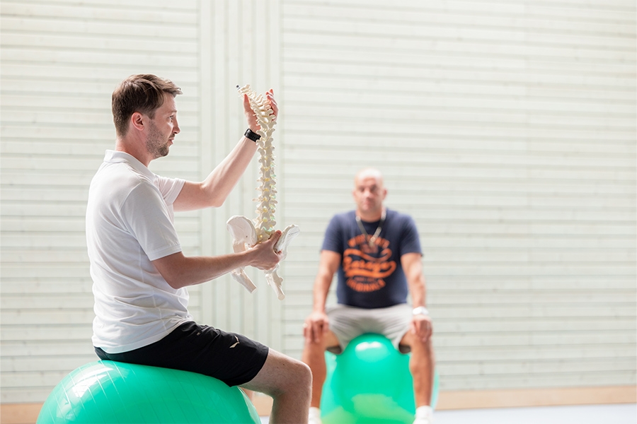 Auf Gymnastikbällen sitzend erklärt ein Therapeut seinen Patienten anhand eines Modells die Wirbelsäule.